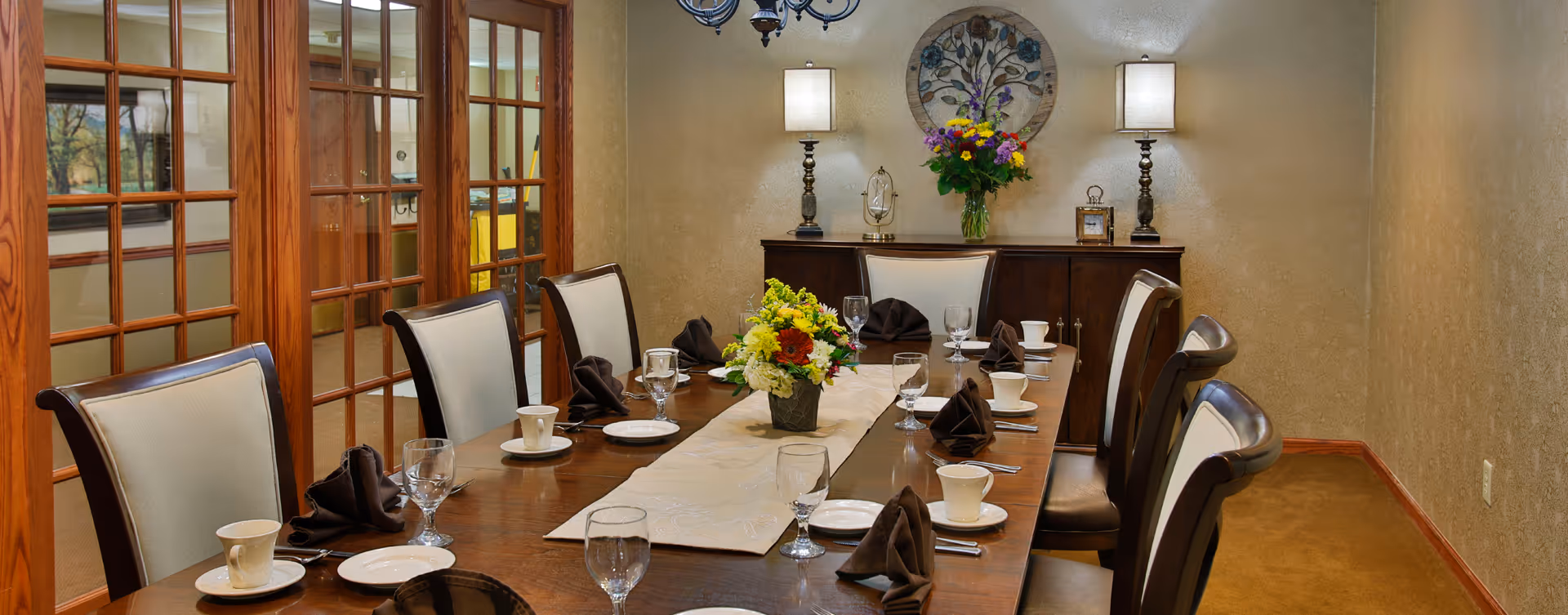 A formal dining room with a long wooden table set for eight people. The table is decorated with a beige runner, two floral centerpieces, folded brown napkins, white cups and saucers, plates, and glassware. The room has beige textured walls, a wooden sideboard with two lamps, a decorative wall hanging, and a chandelier above the table. Wooden framed glass doors are visible on the left side.