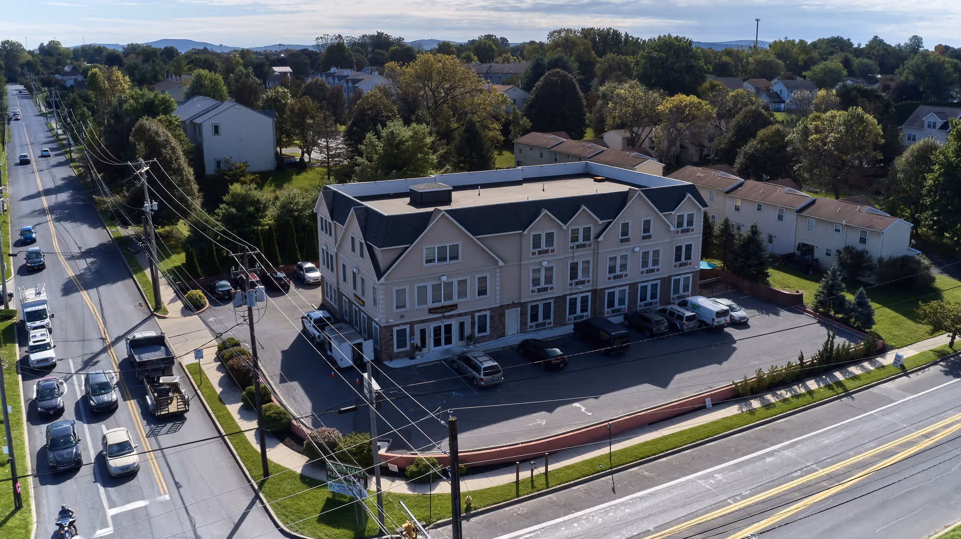 Aerial view of Alexandria Manor showing the building's exterior, parking area, and surrounding landscape with trees and nearby houses.