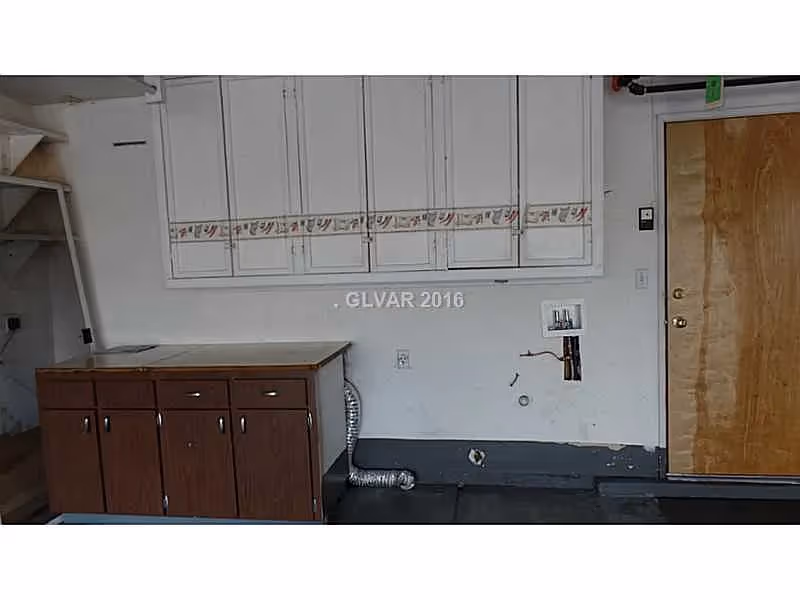 An indoor utility or laundry area with a wooden cabinet on the left, white upper cabinets with decorative trim, a wooden door on the right, and exposed plumbing and ventilation ducts on the wall.