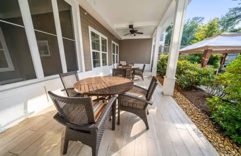 Covered outdoor patio with a round wooden table, four wicker chairs, a ceiling fan, and a landscaped garden with a gazebo visible.