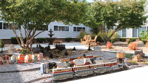 Outdoor garden area with miniature model buildings and a model train set displayed on gravel. There are trees providing shade, a water fountain in the background, and patio chairs near the building with large windows.