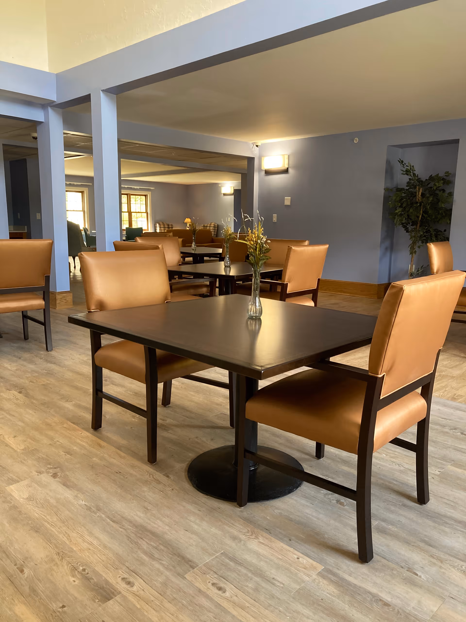 Interior view of a senior living facility dining area with several square dark wood tables, each surrounded by tan cushioned chairs. Each table has a small glass vase with yellow flowers. The room has light wood flooring, blue walls, and soft lighting fixtures on the walls.
