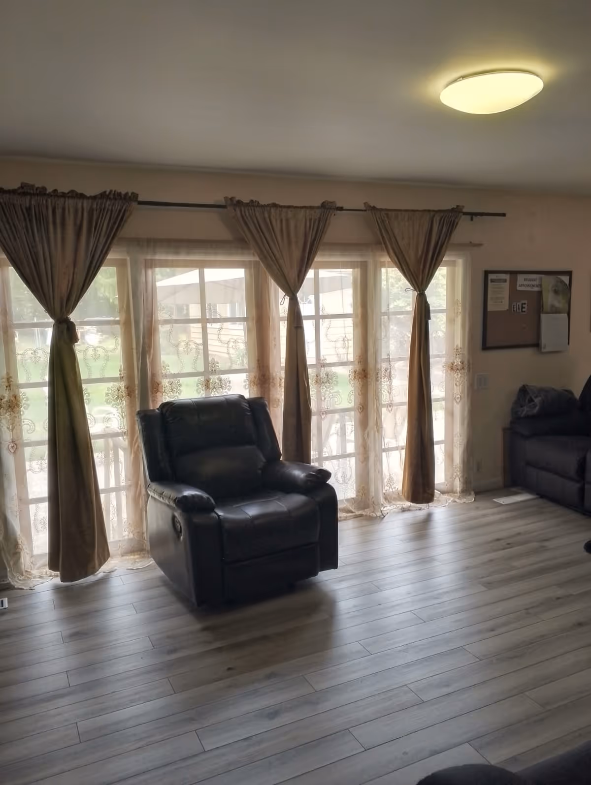 Sunlit living room with a black leather recliner in front of large curtained windows and wood-look flooring.