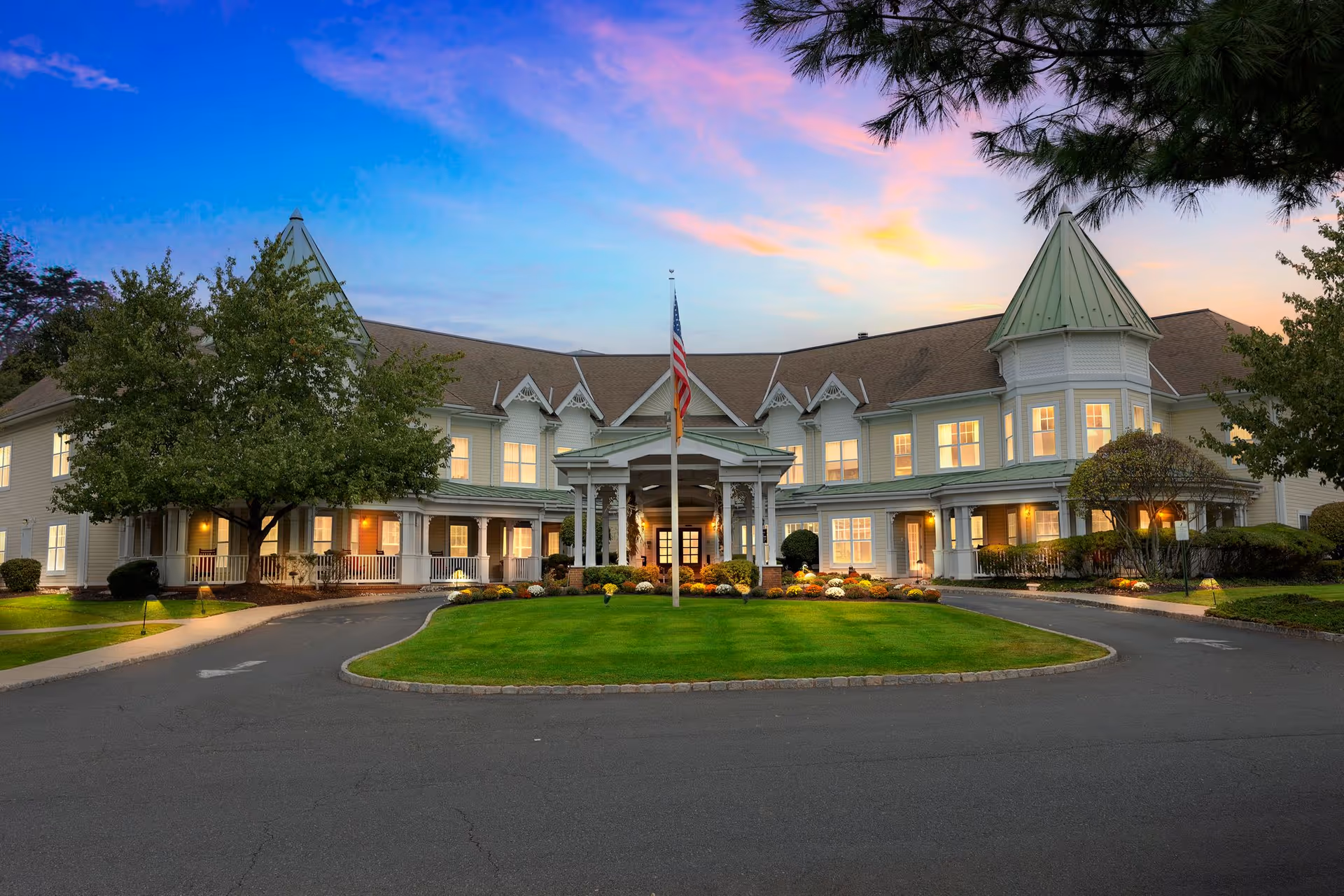 Front exterior view of a large senior living facility building at sunset with a circular driveway, well-maintained lawn, American flag on a flagpole, and illuminated windows.