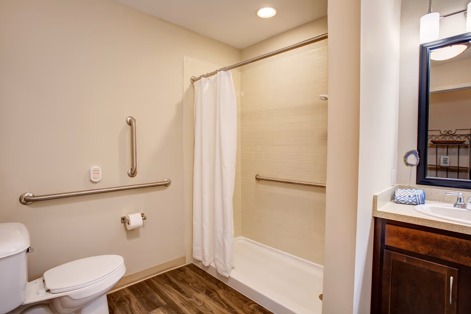 A clean and accessible bathroom featuring a white toilet with grab bars on the wall beside it, a walk-in shower with a white curtain and grab bar, and a vanity with a sink, countertop, and mirror. The floor has wood-like flooring and the walls are painted beige.