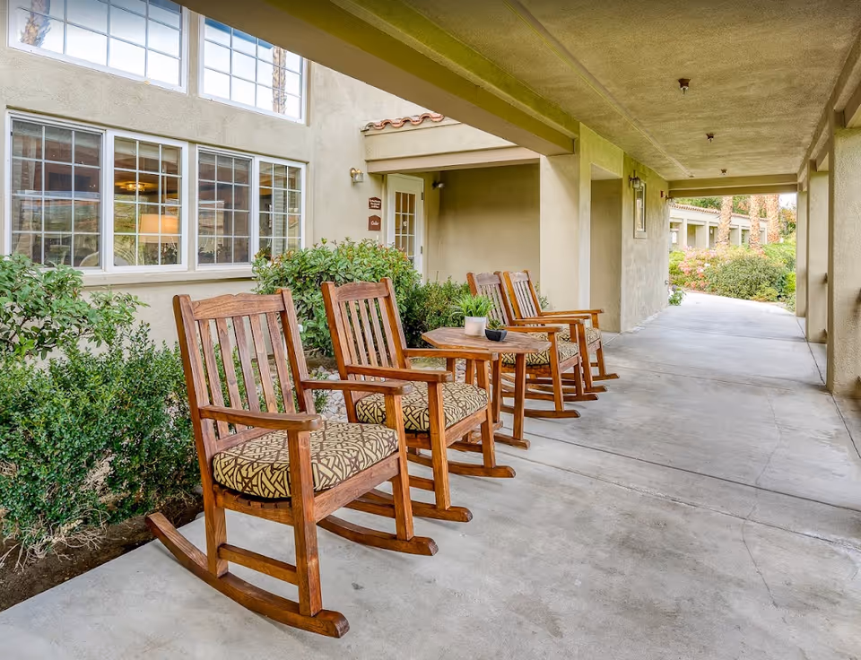 Covered outdoor walkway with wooden rocking chairs and a small table beside shrubs and windows at a senior living facility.