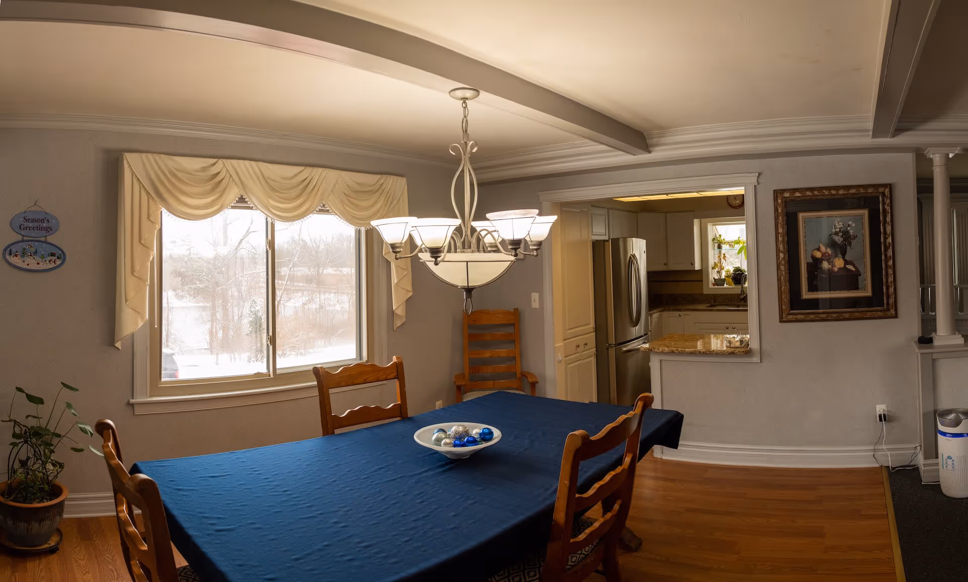 Dining room with a wooden table covered by a dark blue tablecloth and four wooden chairs. A bowl with decorative balls is placed on the table. A chandelier with multiple lights hangs above the table. There is a large window with cream-colored drapes showing a snowy outdoor scene. To the right, there is an open pass-through window to a kitchen with white cabinets and a stainless steel refrigerator. A framed painting hangs on the wall next to the pass-through. A potted plant is on the floor near the window, and a small sign on the wall reads 'Season's Greetings.'