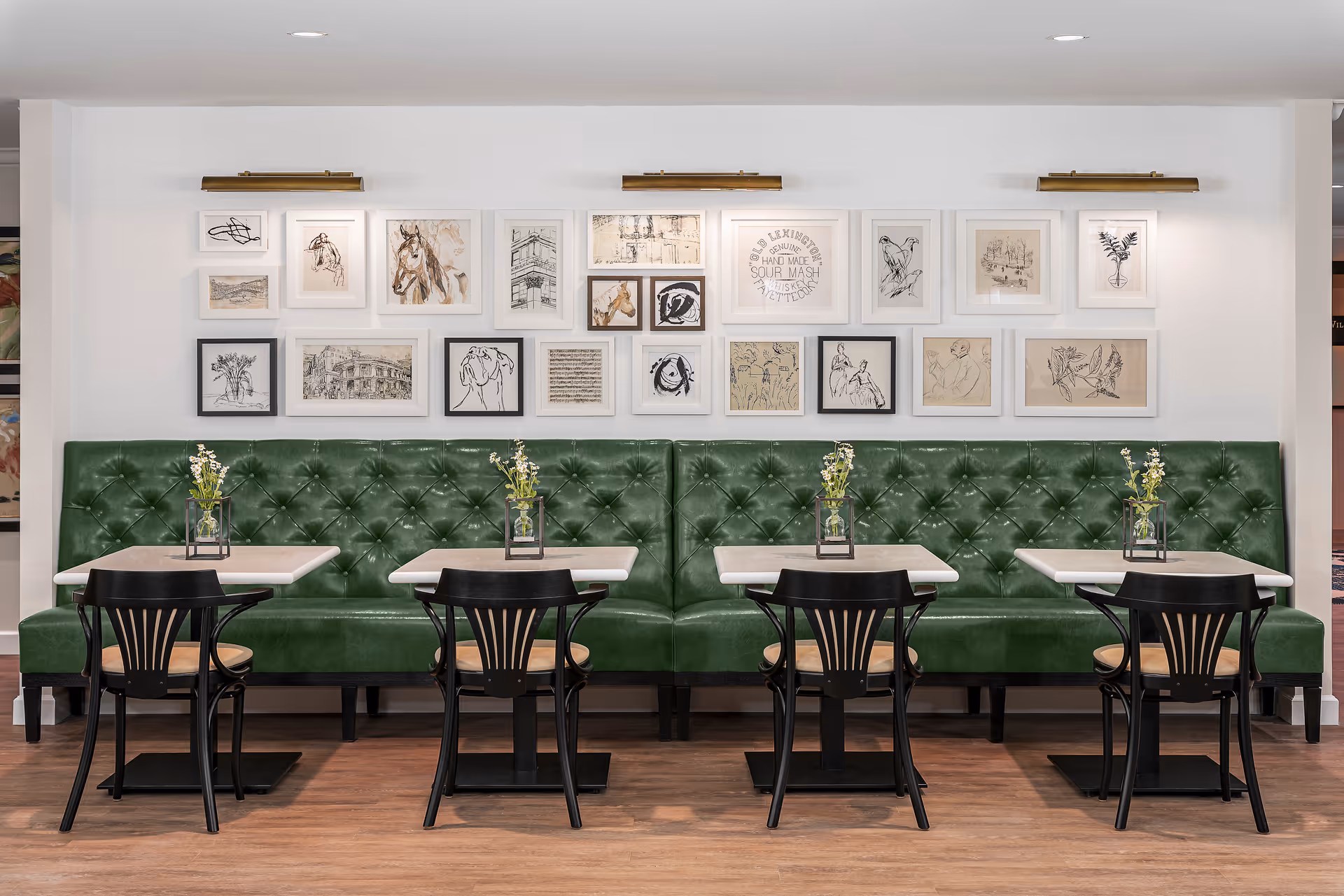 A dining area with a long green tufted bench along the wall, four small square tables each with a black chair, and small flower arrangements in glass containers on each table. Above the bench is a gallery wall with multiple framed sketches and artwork, illuminated by three brass picture lights.