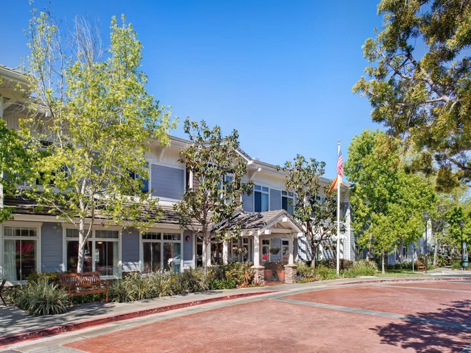 Exterior view of a two-story senior living facility building with a covered entrance, surrounded by trees and landscaping under a clear blue sky. There is a bench on the sidewalk and an American flag on a flagpole near the entrance.