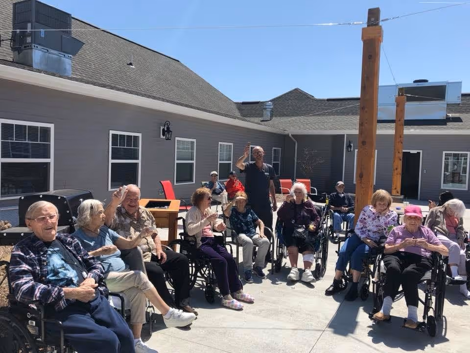 A group of elderly people sitting outside in wheelchairs and chairs in a courtyard area of a retirement living facility. They are enjoying the sunny weather, some are smiling and waving. The building exterior is visible in the background with windows and a clear blue sky overhead.