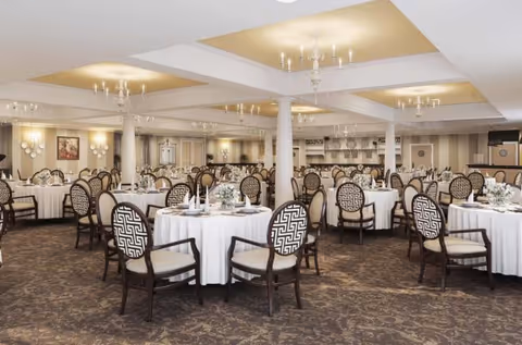 A spacious dining room with multiple round tables covered in white tablecloths, each surrounded by elegant chairs with patterned backs. The room features a carpeted floor, white columns, chandeliers hanging from a coffered ceiling with recessed lighting, and a serving area in the background.