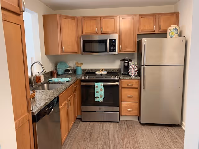 A compact kitchen with wooden cabinets, granite countertops, and stainless steel appliances including a refrigerator, oven, microwave, and dishwasher. There is a sink with a faucet on the left side, a coffee maker with pods on the counter next to the refrigerator, and a dish rack with plates near the window.