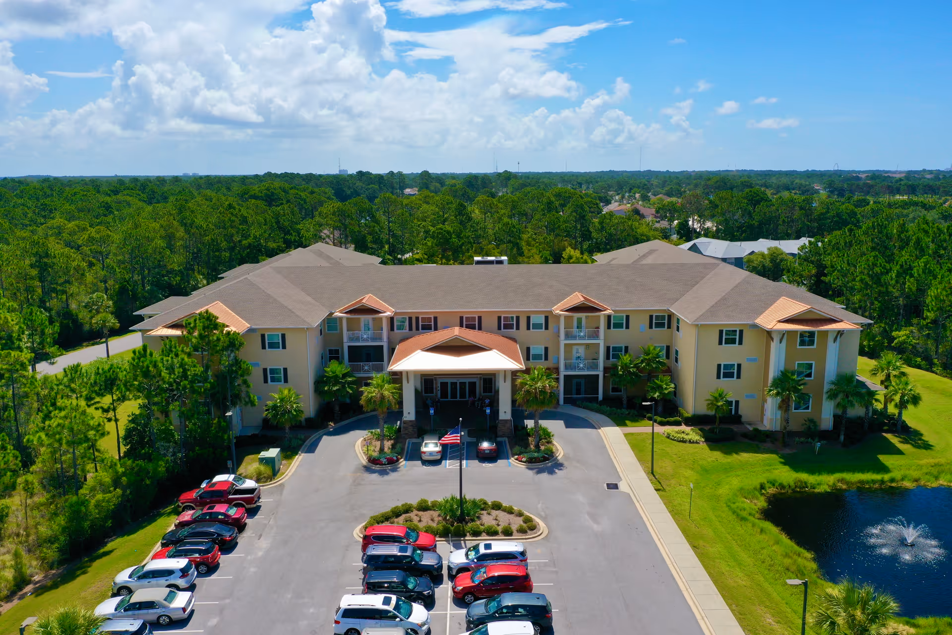 Aerial view of a large, two-story senior living facility building with a beige exterior and brown roof, surrounded by green trees and landscaping. There is a parking lot with several cars in front of the building and a small pond with a fountain to the right. The sky is partly cloudy with blue patches.