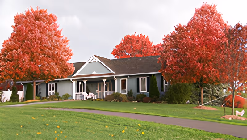 Single-story building with a porch surrounded by vibrant red-leaved trees and a well-maintained green lawn under a cloudy sky.