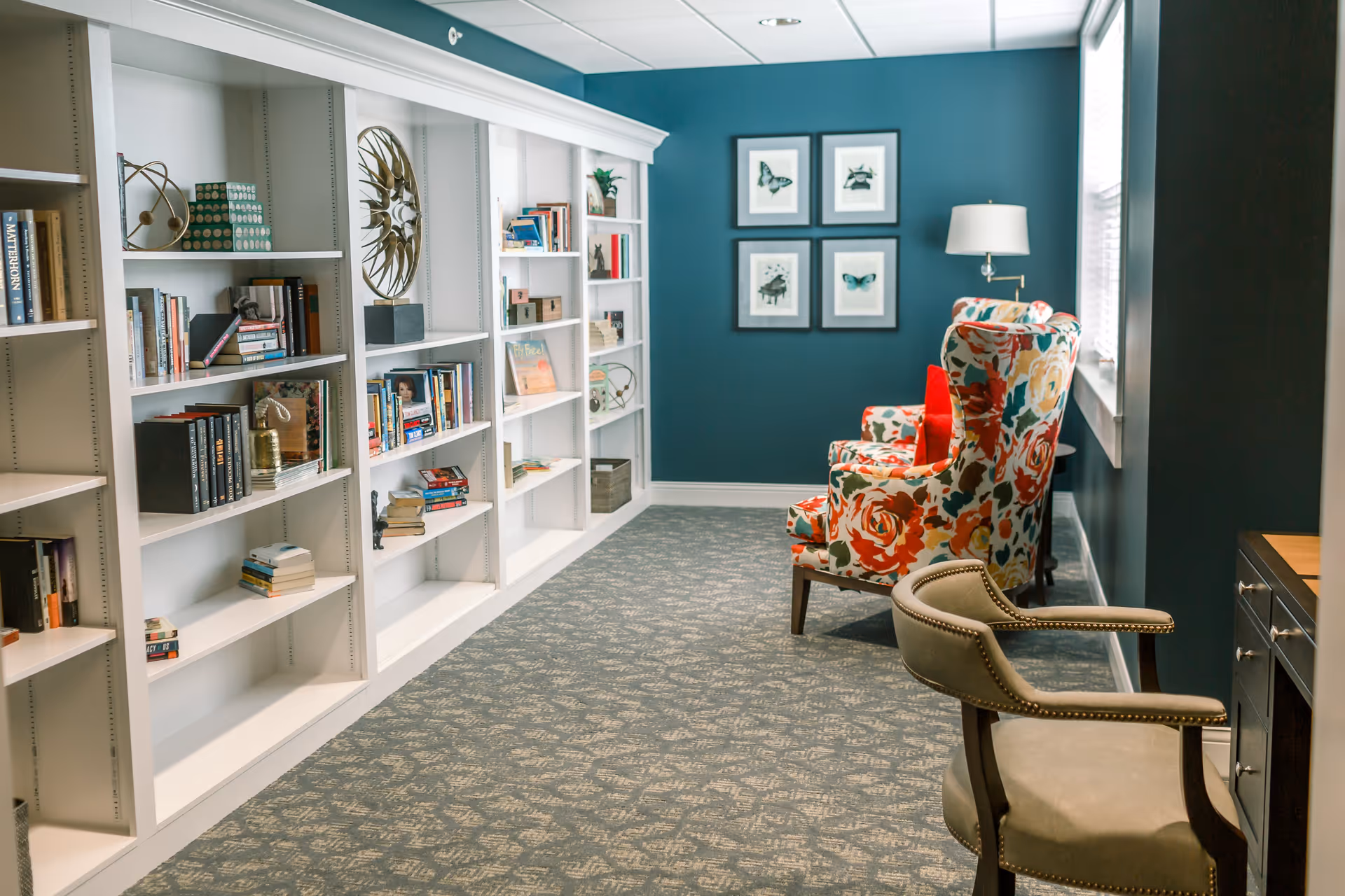 A cozy reading or sitting area in a senior living facility featuring white built-in bookshelves filled with books and decorative items along one wall. The opposite wall is painted teal and adorned with four framed butterfly prints. There are two floral-patterned armchairs with red cushions near a window, accompanied by a floor lamp, and a beige upholstered chair with wooden arms near a dark wooden desk.