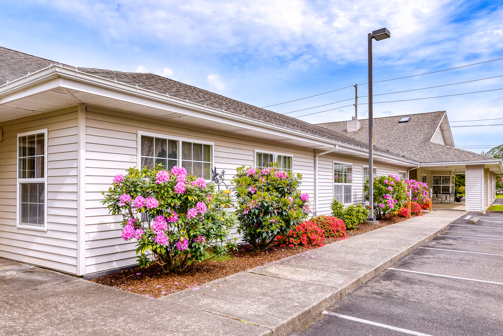 Exterior view of a single-story building with beige siding, several windows, and a covered entrance. There are pink and red flowering bushes planted along the sidewalk next to a parking lot under a partly cloudy sky.
