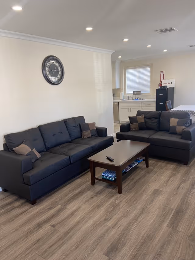 A living room area with two dark gray sofas facing each other, each with brown and beige throw pillows. A wooden coffee table with a remote control on top is placed between the sofas. The room has wood-look flooring and white walls with a large round clock hanging on one wall. In the background, there is a kitchen area with white cabinets, a window with blinds, and a black filing cabinet with a first aid kit on top.