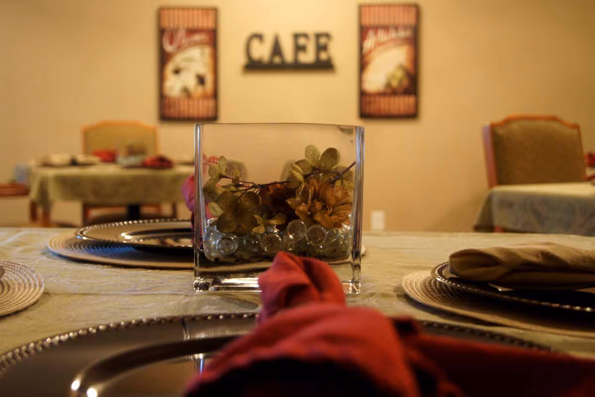 Close-up of a dining table set with a decorative glass vase containing flowers and glass beads, with plates and folded napkins on placemats. In the background, there are more tables and chairs, and a wall decoration with the word 'CAFE' and two framed pictures.