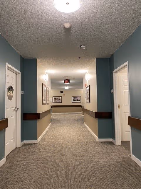 A well-lit hallway in a senior living facility with beige carpet and walls painted in beige and teal. There are white doors on both sides of the hallway, some with decorations. Handrails run along the walls, and framed pictures are hung on the walls at the end of the hallway. Ceiling lights illuminate the corridor, and an exit sign is visible in the distance.