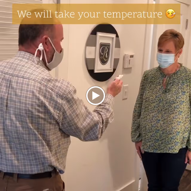 A man wearing a face mask is using a handheld device to take the temperature of a woman who is also wearing a face mask. They are standing indoors near a wall with a decorative round frame and a thermostat.