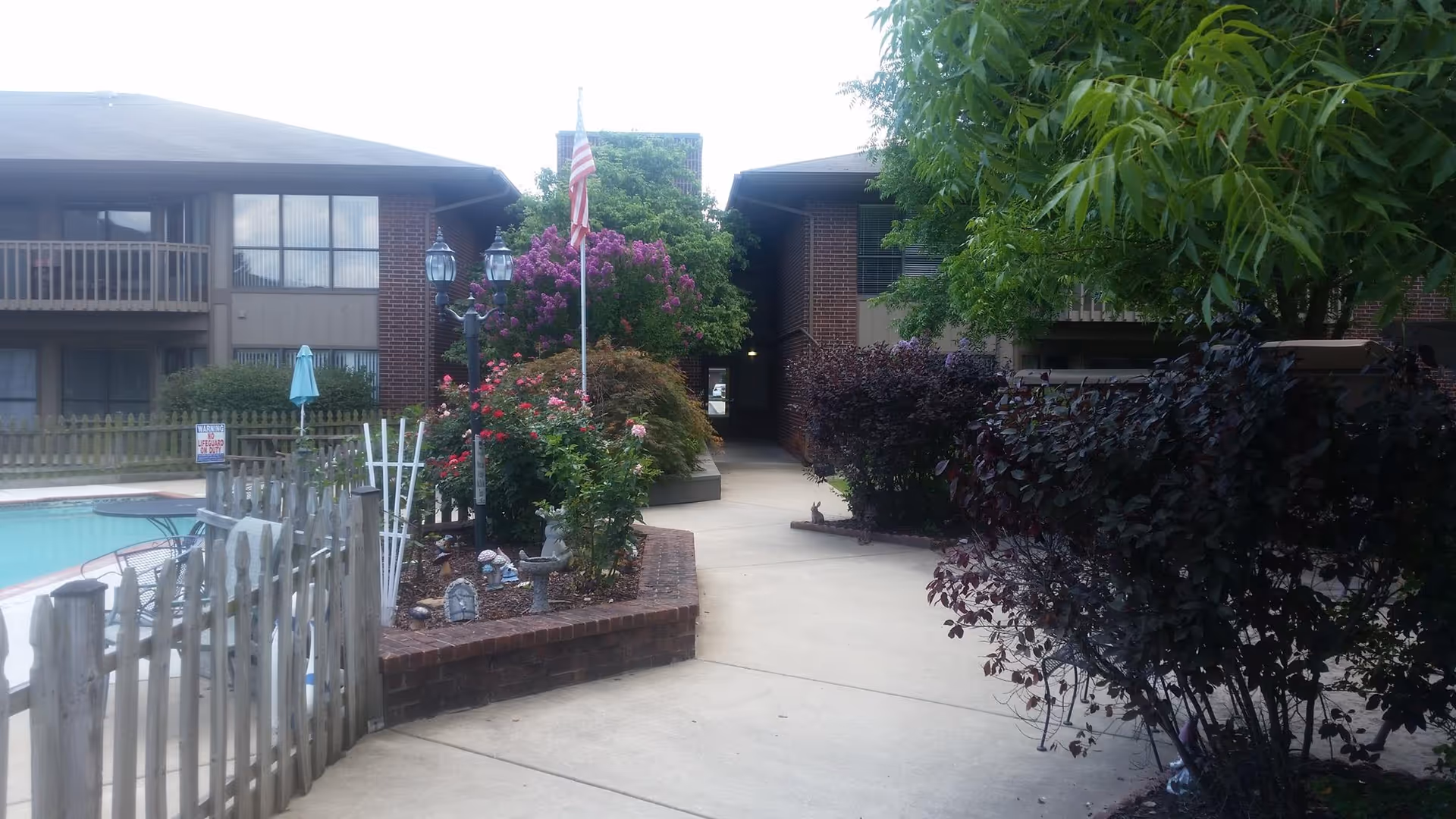 Outdoor view of a senior living facility showing a paved walkway surrounded by lush green trees and colorful flowering bushes. To the left, there is a fenced swimming pool area with patio furniture and an umbrella. The building has brick walls and large windows, with an American flag on a pole near the center.