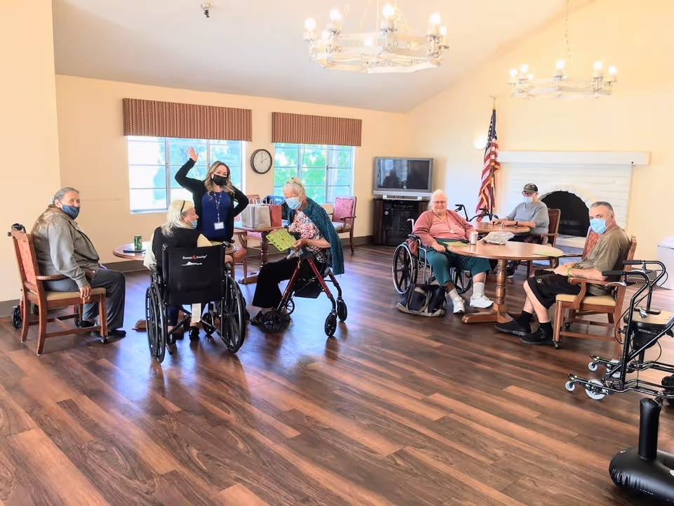 Residents and a staff member wearing masks gathered around tables and wheelchairs in a bright common room with wood floors and an American flag.