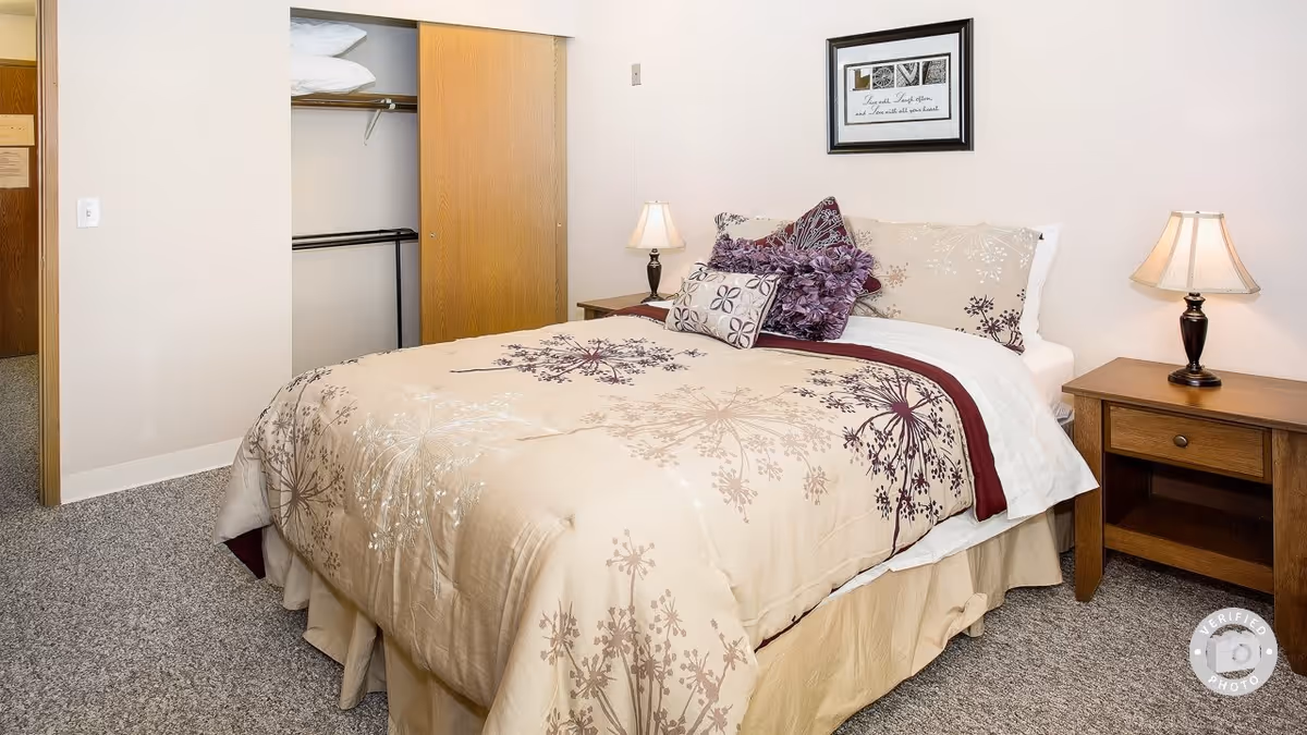 A bedroom with a bed covered in beige and purple floral bedding, two wooden nightstands each with a lamp, an open closet with sliding wooden doors, and a framed picture on the wall above the bed.