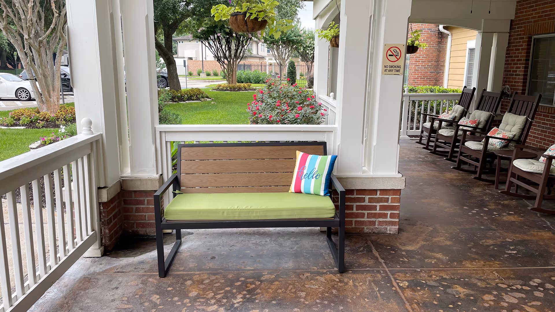 A covered outdoor porch area with a bench featuring a green cushion and a colorful striped pillow with the word 'hello'. To the right, there are several wooden rocking chairs with cushions. The porch overlooks a green lawn with trees, bushes, and flowers. A 'No Smoking At Any Time' sign is visible on one of the porch columns.