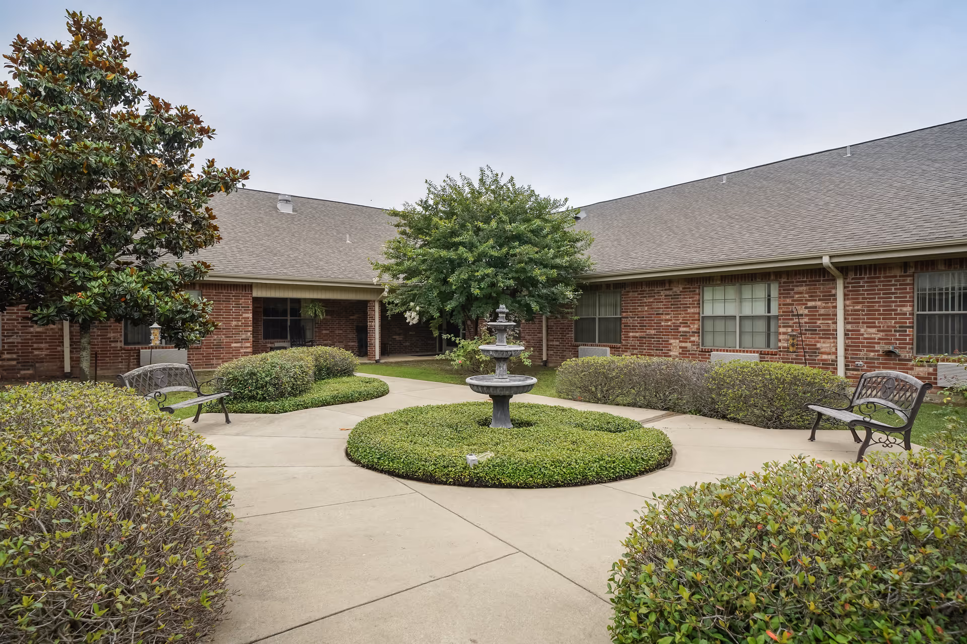 Outdoor courtyard area with a central three-tiered fountain surrounded by neatly trimmed bushes and a concrete walkway. There are benches on either side of the walkway and a brick building with multiple windows in the background under a cloudy sky.