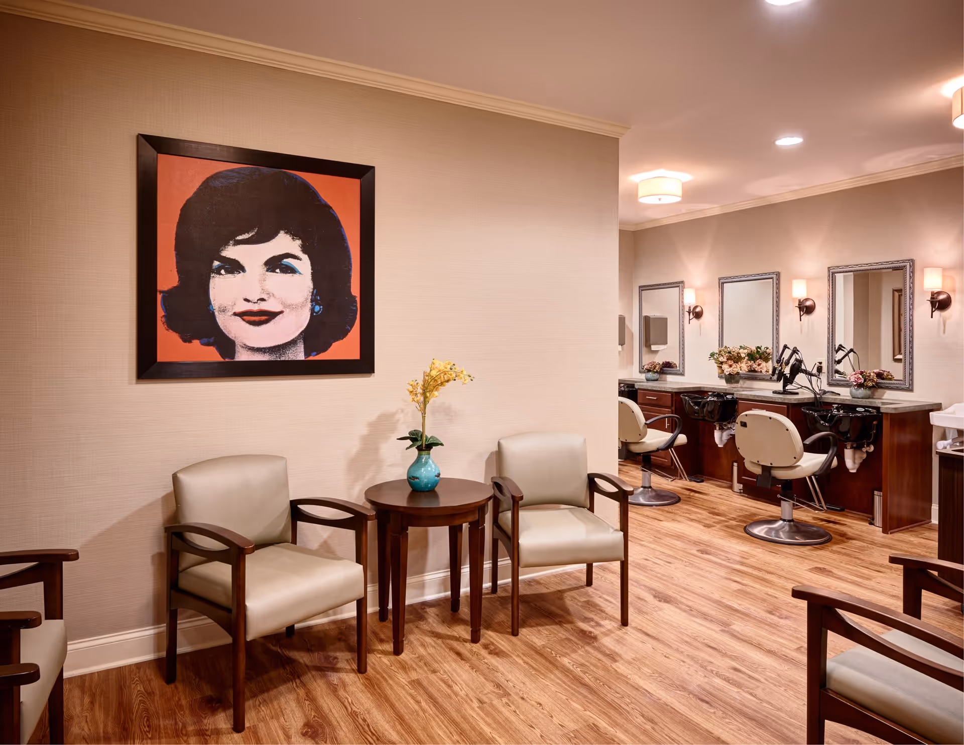 Interior view of a salon area with wooden flooring, beige walls, and ceiling lights. There are two beige cushioned chairs with wooden armrests around a small round wooden table with a blue vase holding yellow flowers. On the wall above the chairs is a framed pop art portrait of a woman with dark hair and red background. In the background, there are three salon stations with mirrors, wall-mounted lights, and salon chairs in front of black wash basins.