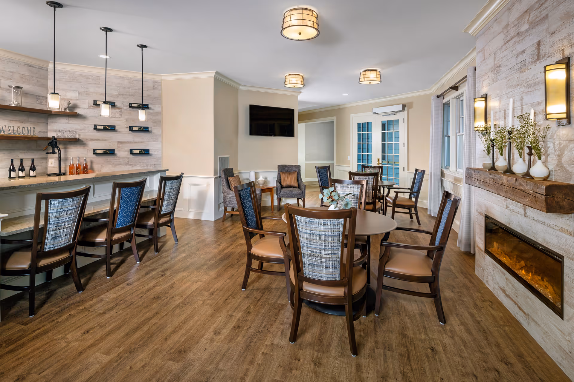 A cozy and well-lit common area with wooden flooring, featuring a round table surrounded by chairs in the center, a bar counter with high chairs on the left, a wall-mounted TV, and a modern fireplace with decorative vases on the right.