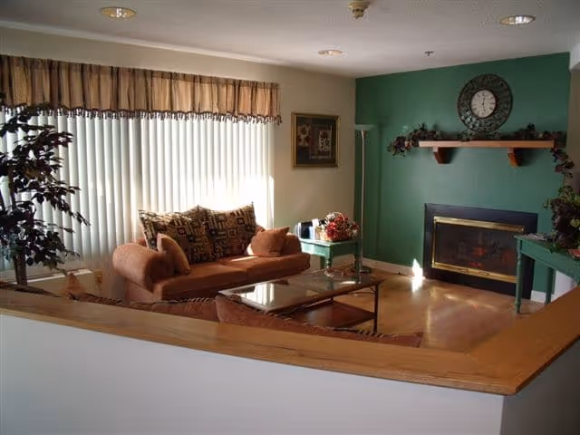 A cozy living room with a brown couch and matching armchair, a glass coffee table, a green side table with a floral arrangement, a green accent wall with a fireplace and a clock above it, and a large window with vertical blinds letting in natural light.