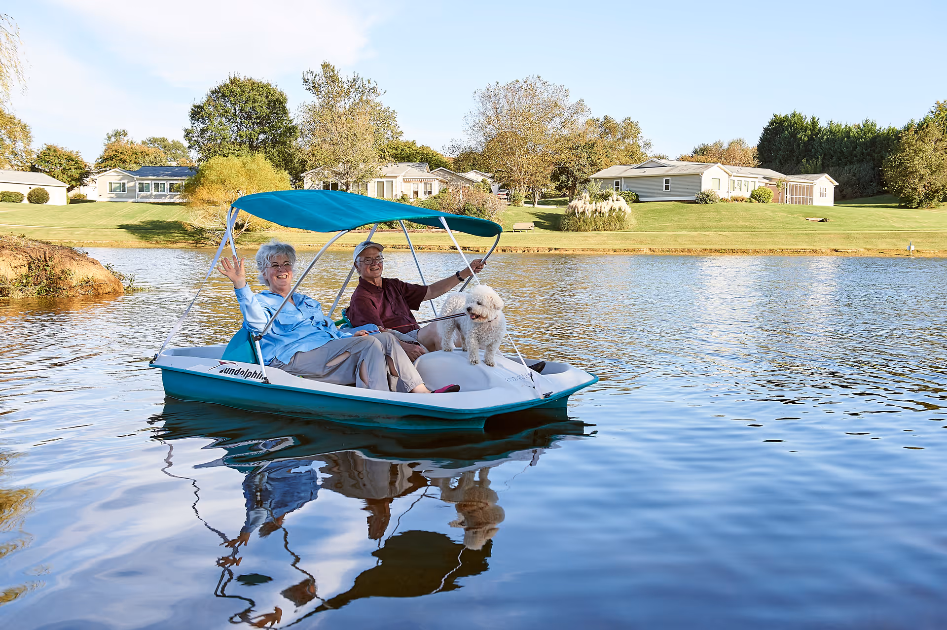 An elderly couple and a small white dog are riding a pedal boat on a calm lake with green lawns and houses in the background under a clear sky.