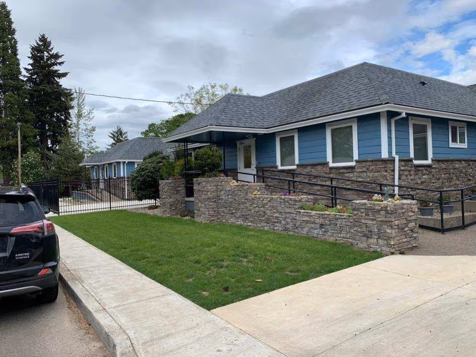 Exterior view of a single-story building with blue siding and stone accents, featuring multiple windows and a sloped roof. There is a paved sidewalk and a green lawn in front, with a black car parked on the street to the left. A black metal fence and gate are visible in the background, along with trees and a cloudy sky.