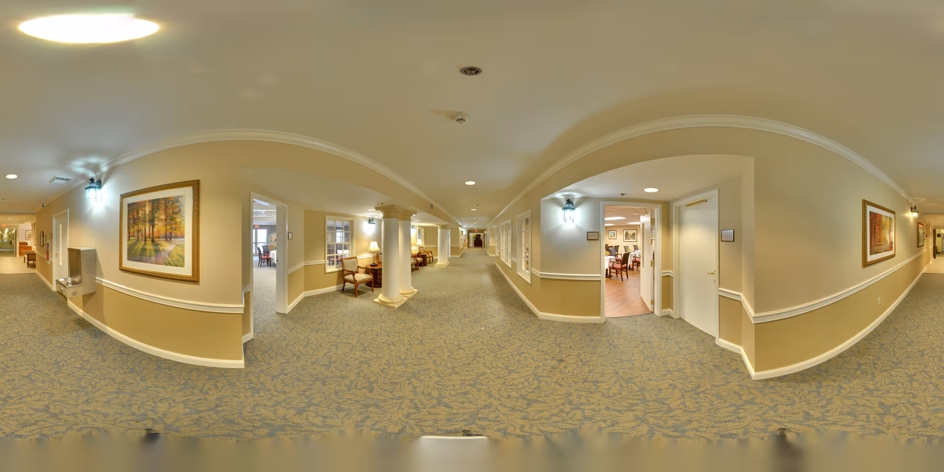 A wide, carpeted hallway in a senior living facility with beige walls and white trim. The hallway features framed artwork on the walls, several wooden chairs with cushions, and columns. There are open doorways leading to rooms with tables and chairs, likely common or dining areas. The ceiling has recessed lighting and wall-mounted lamps providing illumination.