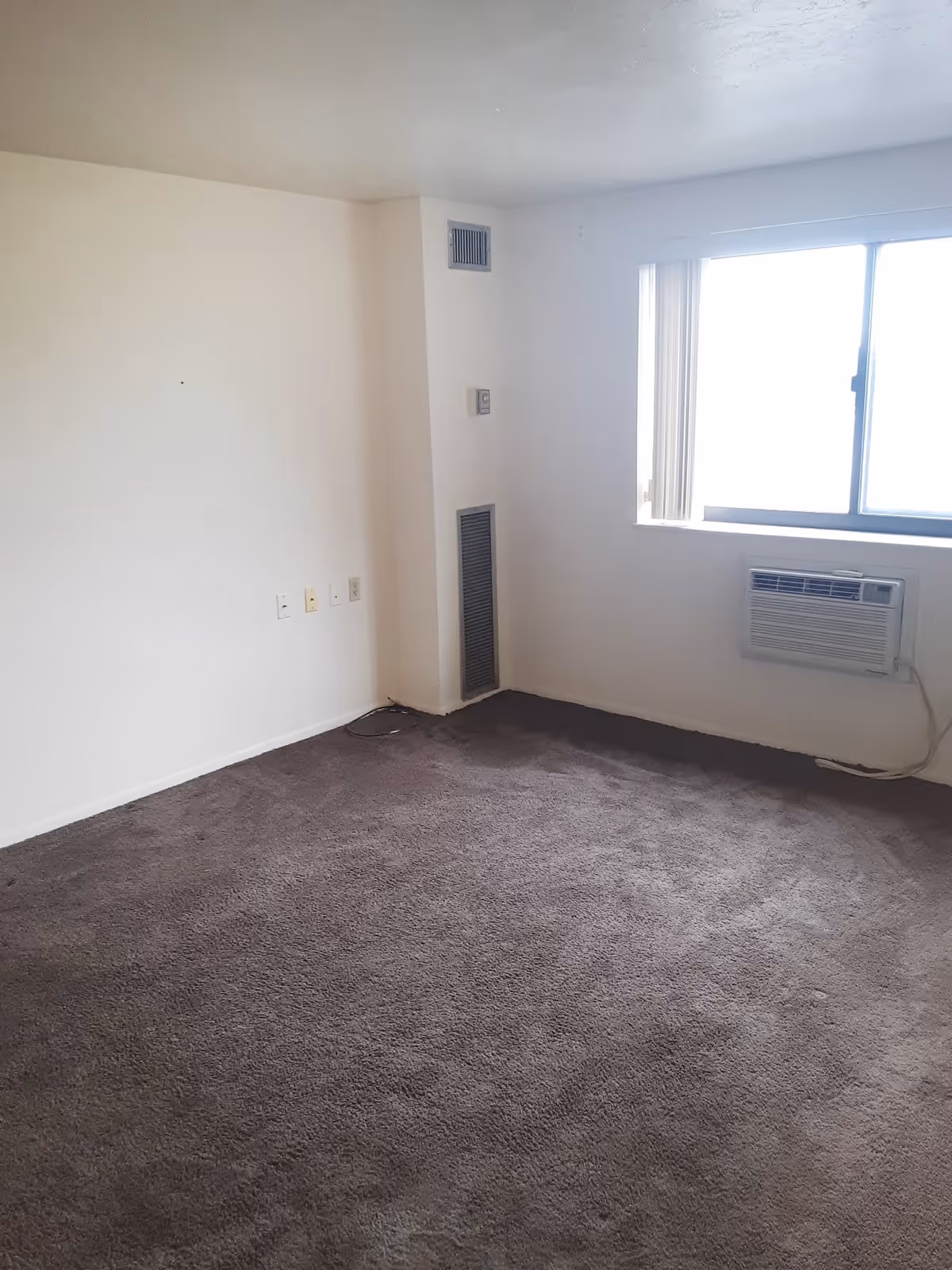 Empty room with beige walls and dark carpet flooring. There is a window with vertical blinds and a wall-mounted air conditioning unit below it. The room has a vent and thermostat on one wall, and several electrical outlets near the floor.