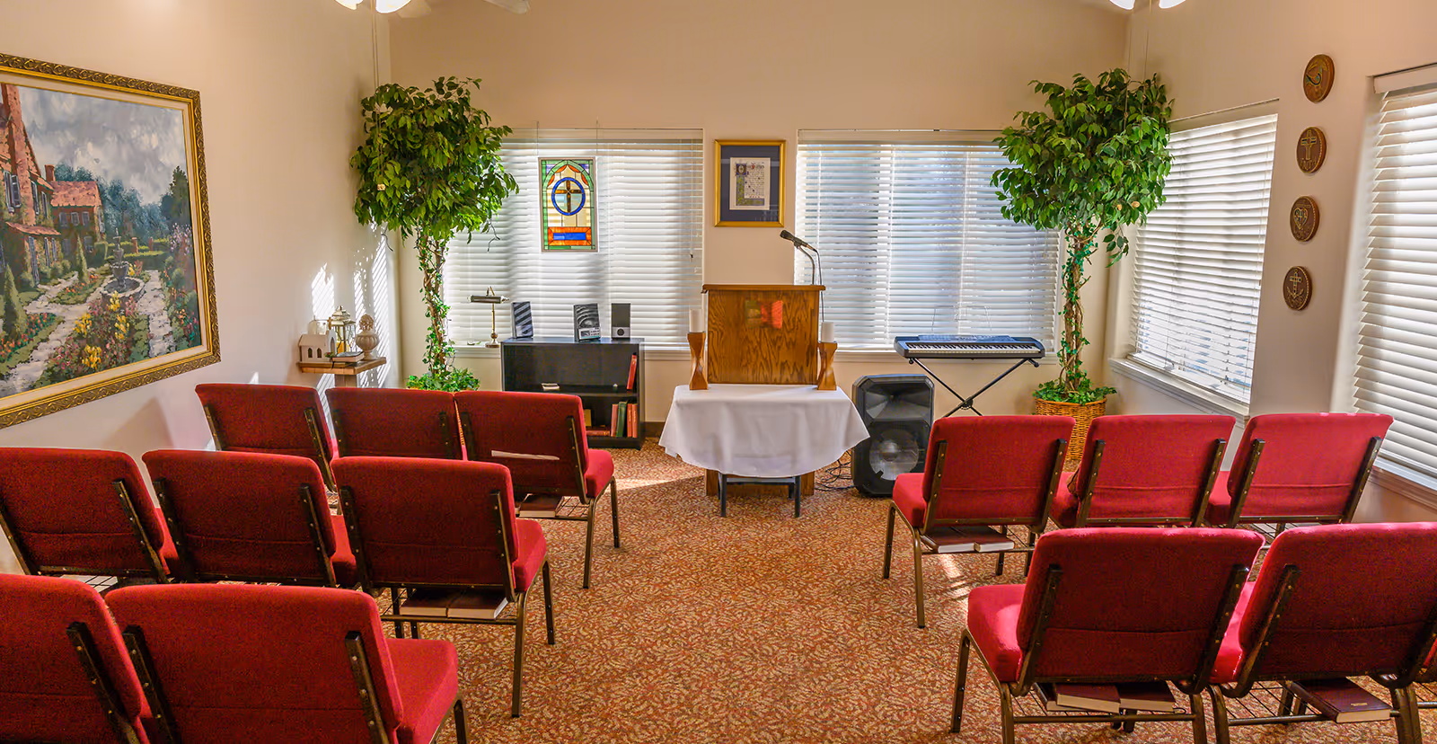 Small chapel room with rows of red cushioned chairs facing a wooden lectern on a table covered with a white cloth. The room has carpeted floor, two large potted plants, a keyboard, a speaker, and windows with blinds. There is a framed painting on the left wall and decorative plaques on the right wall.