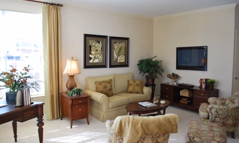 A cozy living room with a beige loveseat adorned with two patterned cushions, a wooden side table with a lamp and small plant, a wooden coffee table with magazines, and a floral armchair with a matching ottoman. The room also features a wooden TV stand with a flat-screen TV mounted on the wall, framed botanical artwork, a large window with beige curtains, and decorative plants.