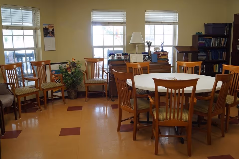 A room with a round white table surrounded by wooden chairs with green cushions. Additional wooden chairs are lined up against the wall with three large windows letting in natural light. There is a piano with framed photos and a lamp on top, a bookshelf filled with books, and a plant in the corner.