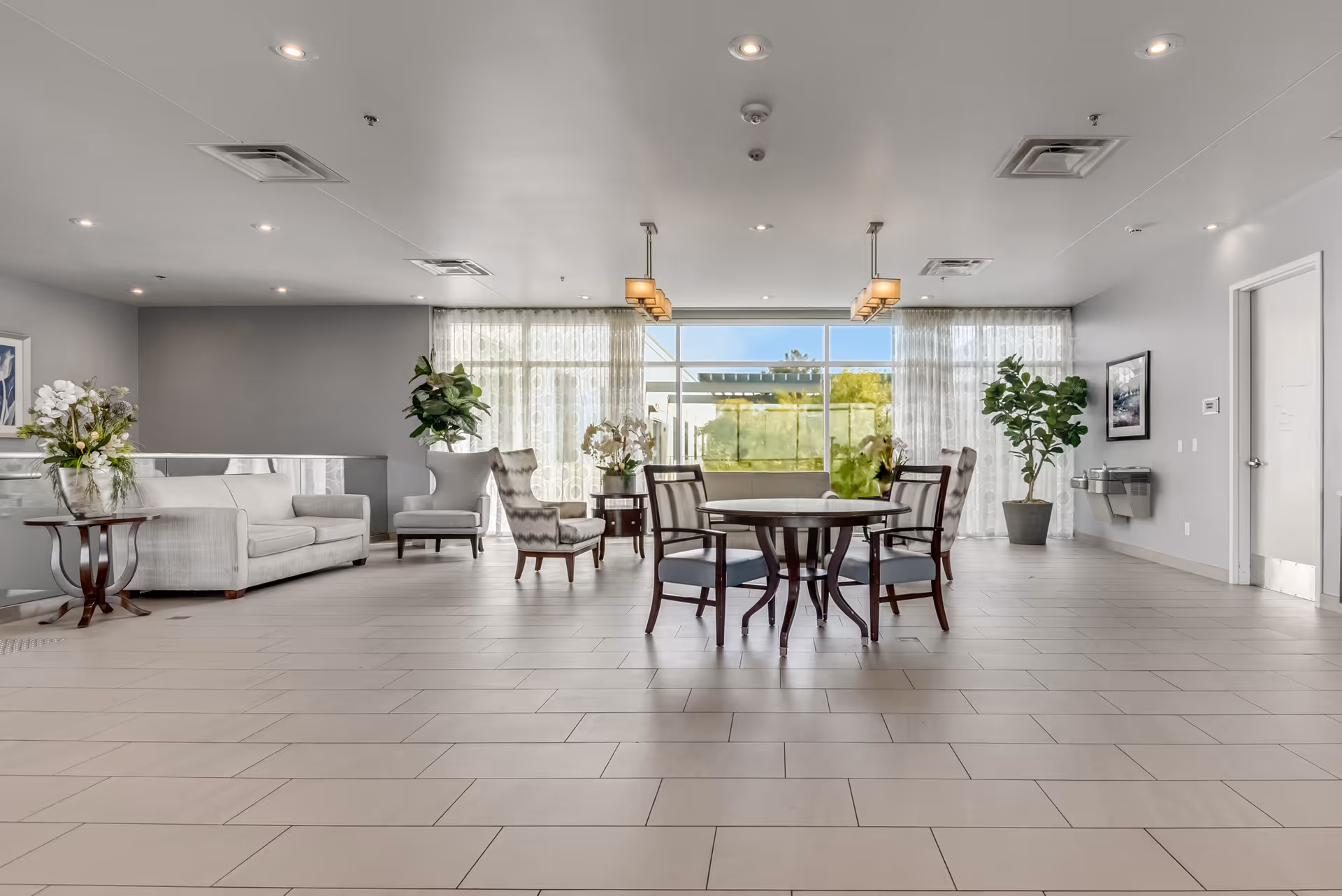 A spacious and well-lit common area with large windows covered by sheer curtains. The room features a round table with four chairs in the center, a light gray sofa, two armchairs, side tables with flower arrangements, and potted plants. The floor is tiled, and the walls are painted light gray with framed artwork. Ceiling lights and two pendant lights hang from the ceiling.