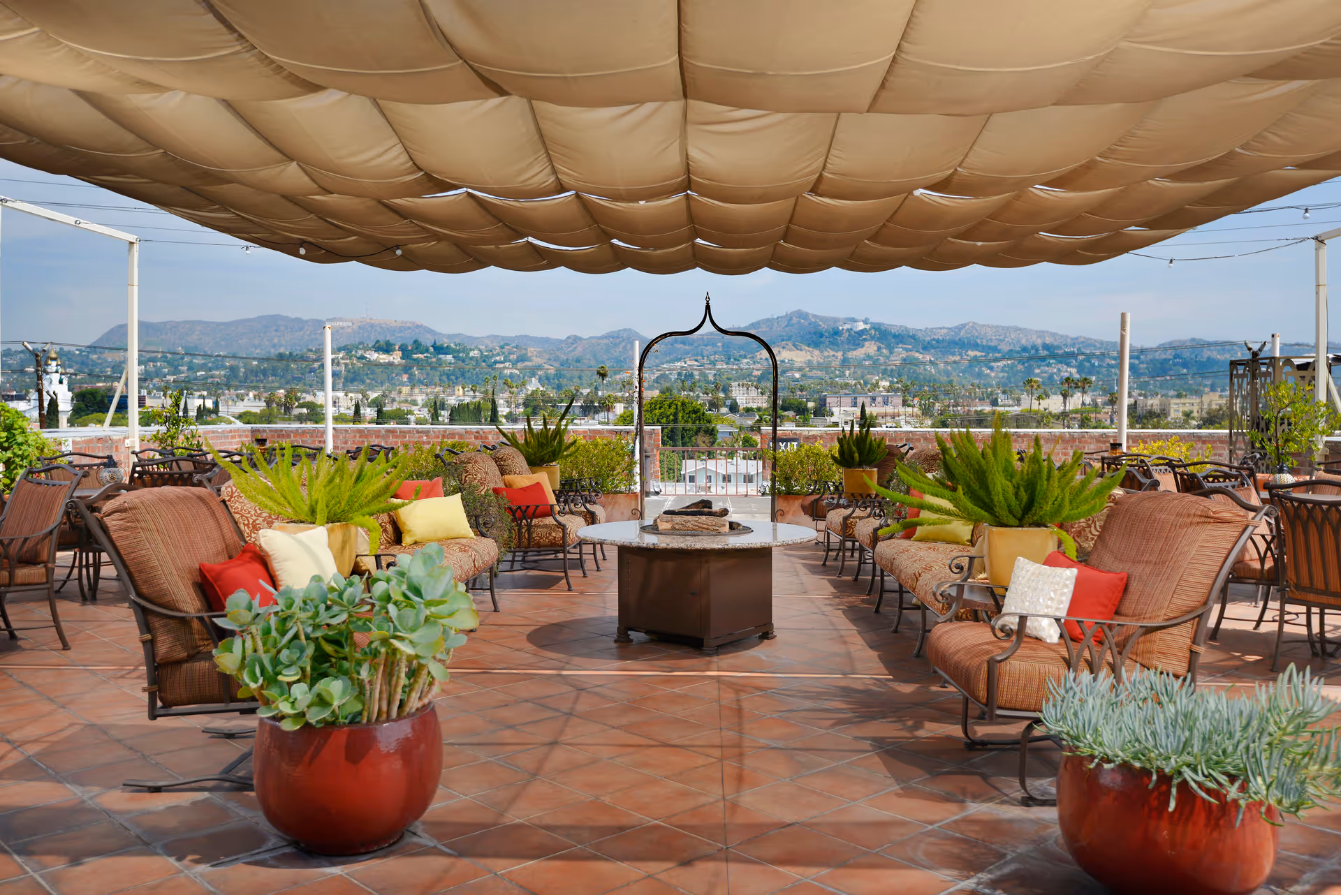 Outdoor patio area with cushioned seating, colorful pillows, potted plants, and a central fire pit under a large fabric canopy with a scenic mountain view in the background.