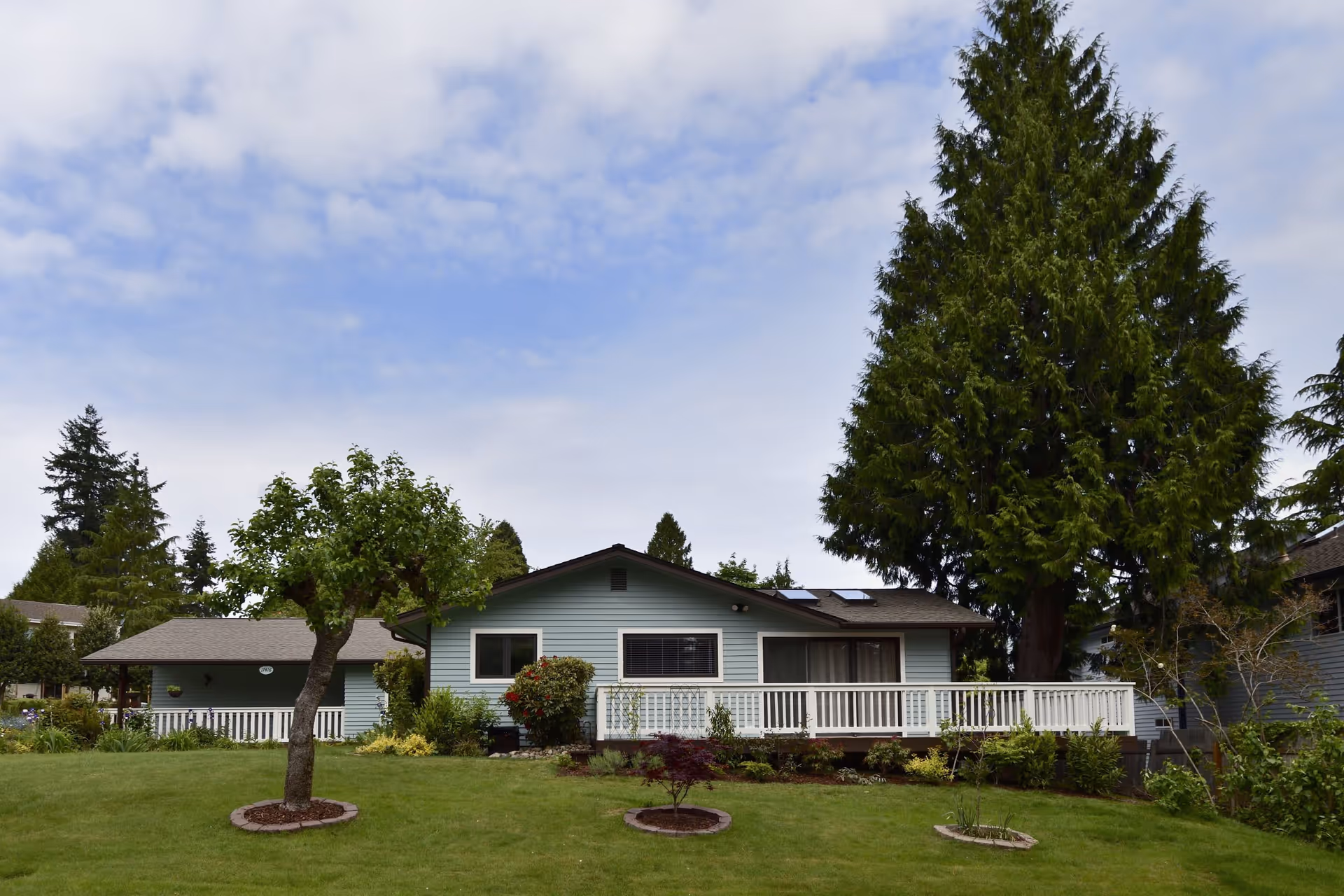 Single-story light blue house with a white railing porch, surrounded by a well-maintained green lawn with small trees and shrubs, under a partly cloudy sky.