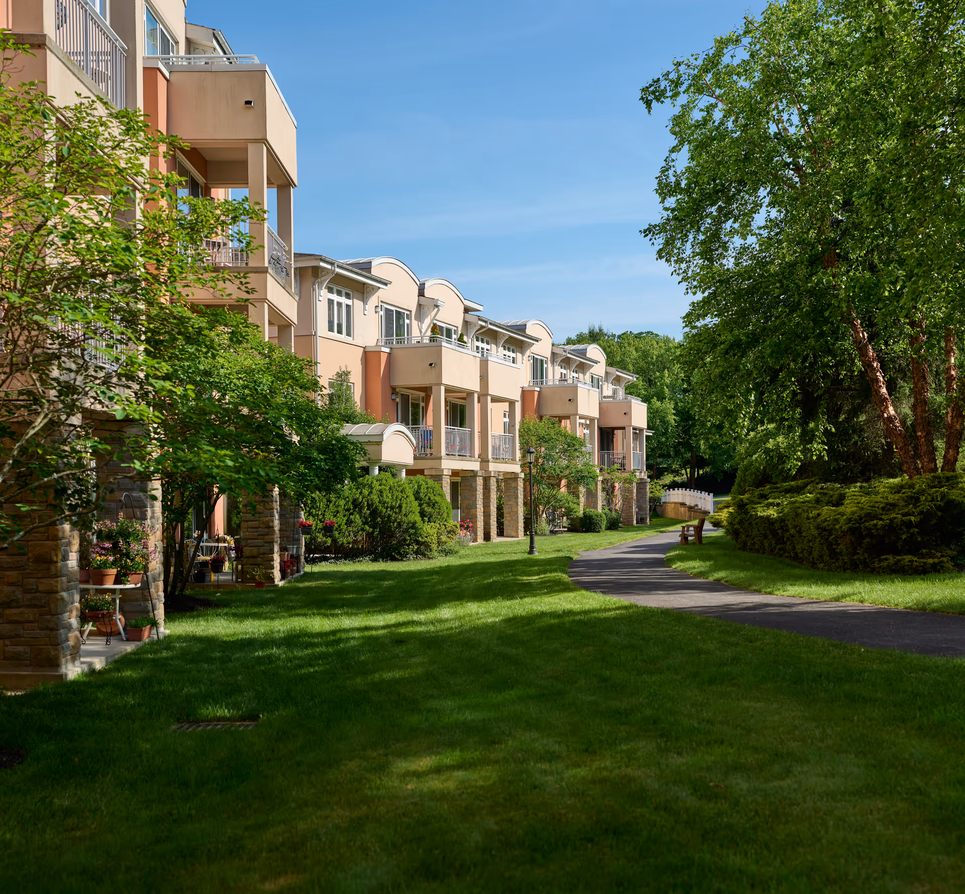 A sunny outdoor view of a senior living facility with a paved walking path, green grass, trees, and a multi-story building with balconies and stone pillars.