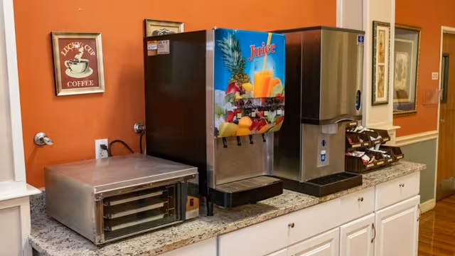 A countertop with a juice dispenser, a coffee machine, a small warming oven, and a rack holding various coffee creamers and sugar packets. The wall behind is painted orange with framed pictures hanging on it.