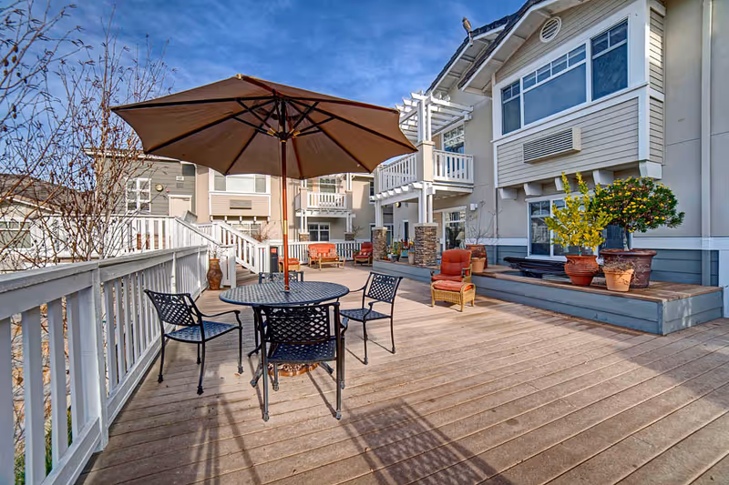 Outdoor patio area at Fremont Hills with a round metal table and four chairs under a large beige umbrella. The deck is made of wooden planks and has white railings. There are potted plants and cushioned chairs along the building wall, which has large windows and balconies. The sky is clear and blue.