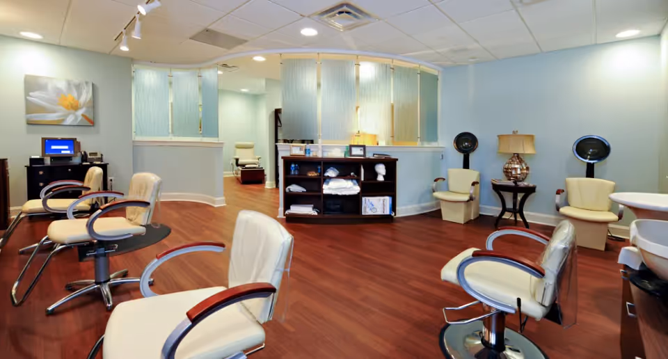 Interior view of a salon area with several cream-colored salon chairs, hair dryers, a small table with a lamp, and a shelving unit with towels and supplies. The room has wooden flooring, light blue walls, and a ceiling with recessed lighting.