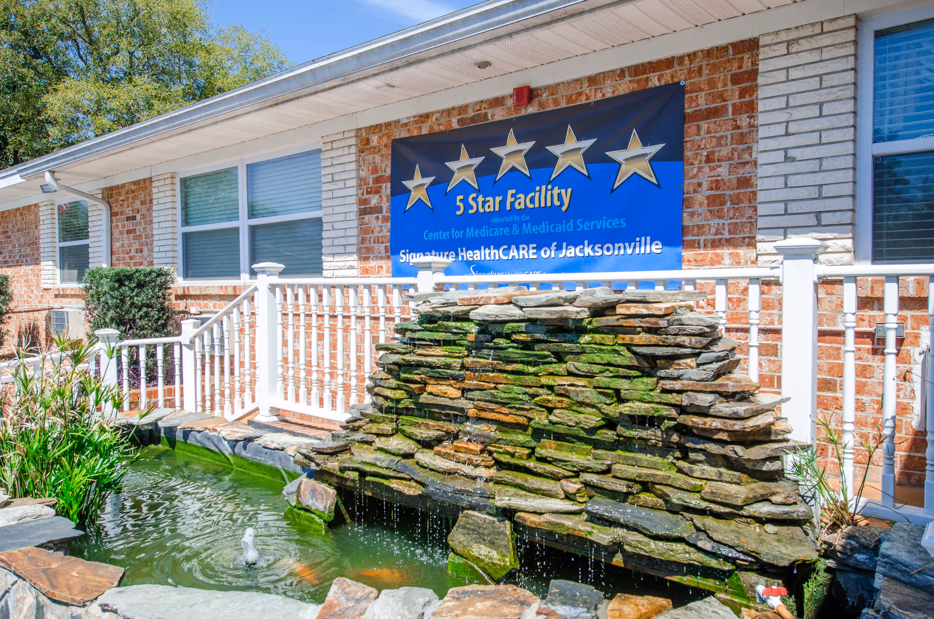 Front exterior of a nursing center with a rock waterfall pond, white railing, and a blue '5 Star Facility' banner on a brick wall.