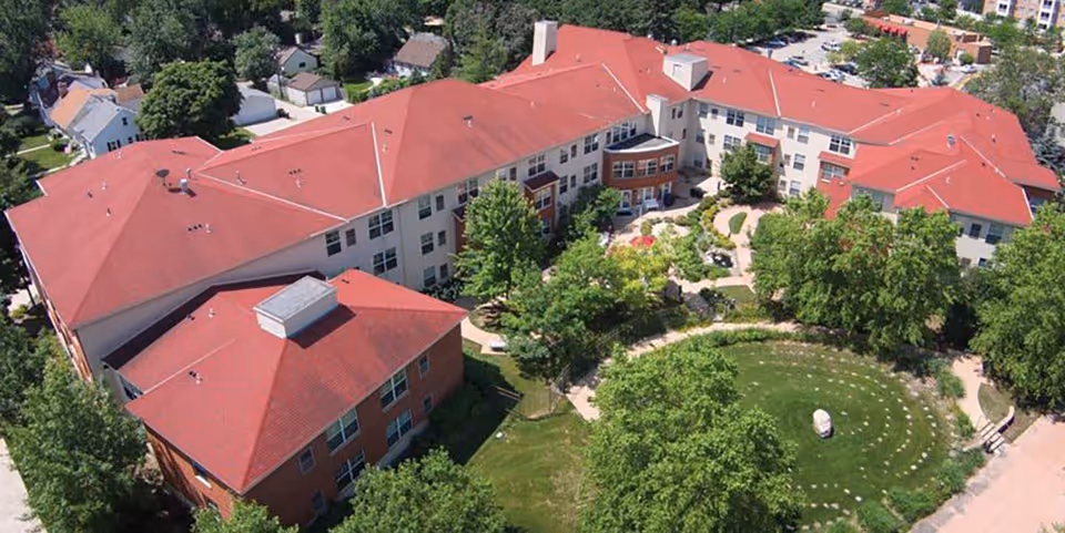 Aerial view of The Pines Senior and Assisted Living facility showing a large building with red roofs surrounded by trees and greenery. There is a circular garden area with pathways and landscaping in front of the building.