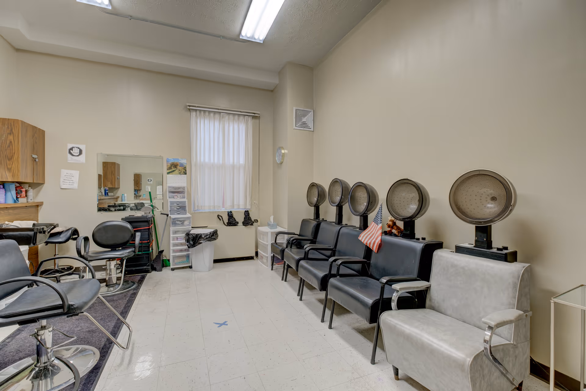 Interior view of a hair salon area with several black salon chairs and hair dryers lined up against the wall. There is a window with sheer curtains, a mirror, and various salon supplies on shelves and carts. The room has beige walls and a tiled floor.