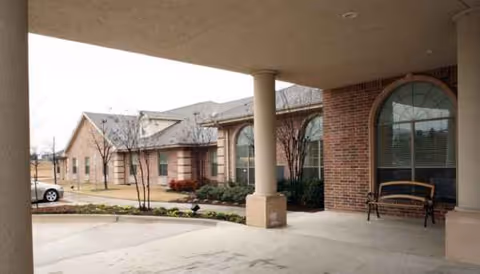 Exterior view of a brick senior care facility with arched windows and a bench.
